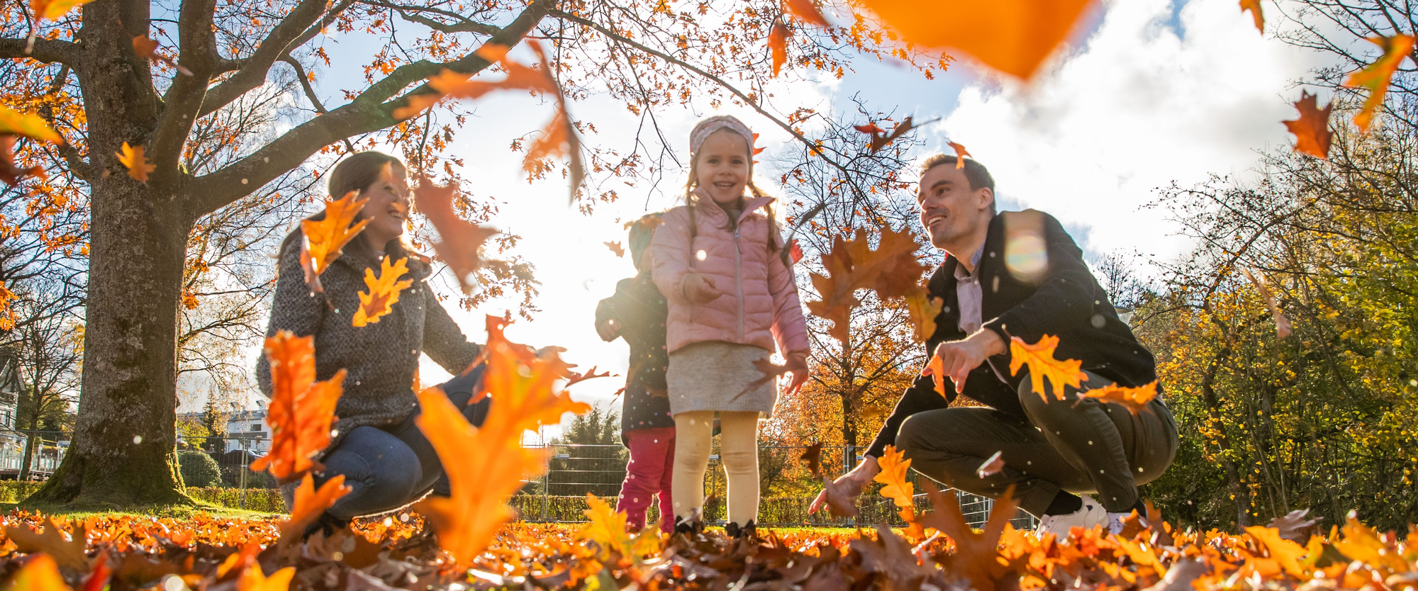 Eine Familie mit zwei Kindern spielt draußen im Sonnenschein und wirft bunte Herbstblätter in die Luft.