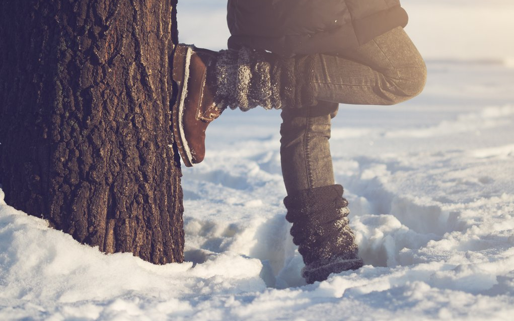 Frau lehnt steht mit dem Rücken zu einem Baum im Schnee und lehnt sich mit einem Fuß an.