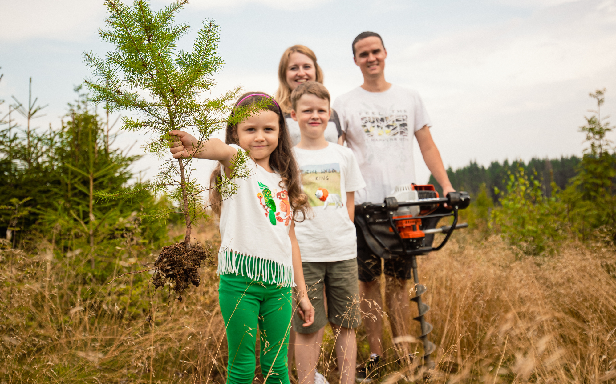 Eine Familie mit zwei Kindern will einen neuen Baum einpflanzen, den das kleine Mädchen noch in der Hand hält.
