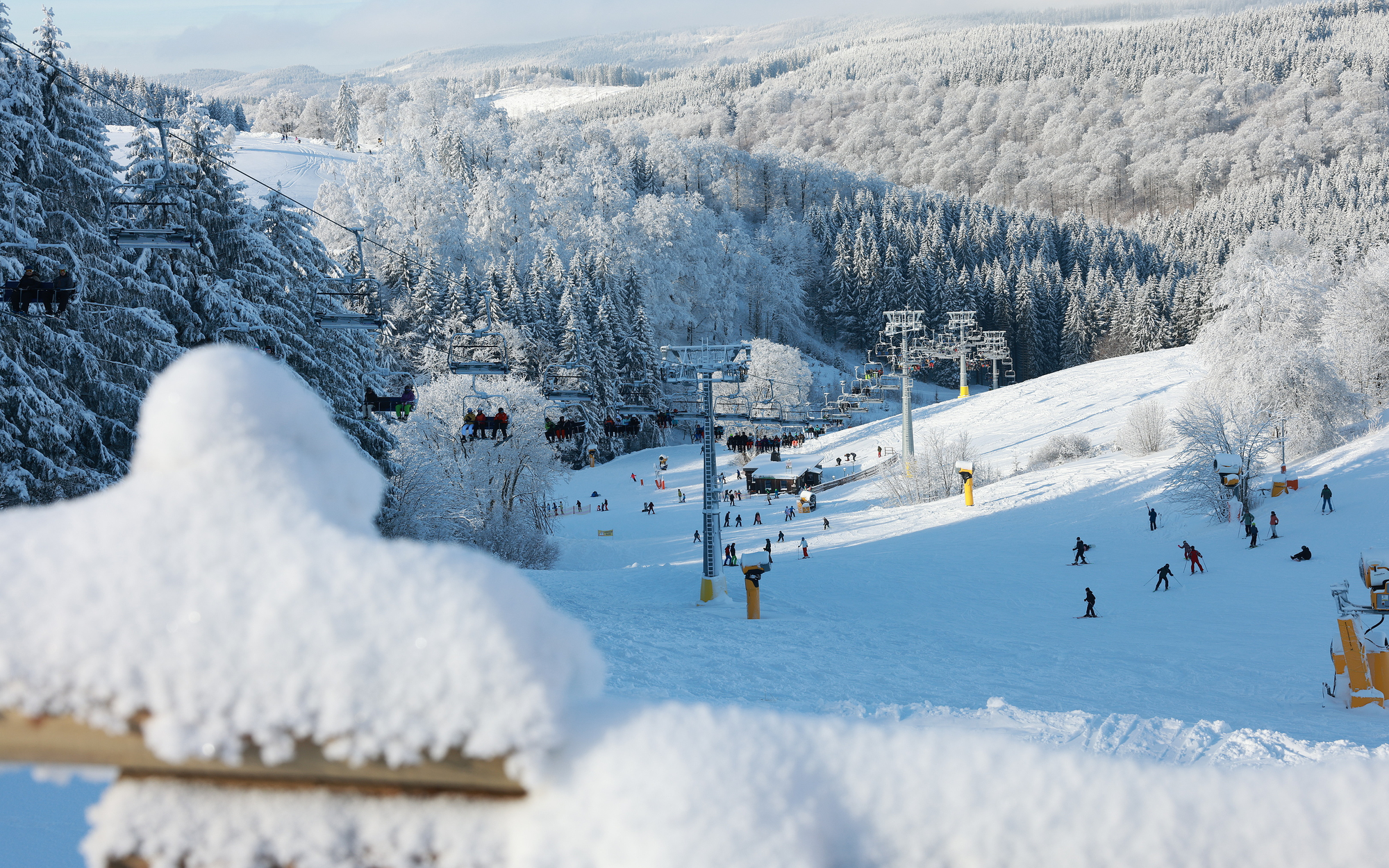 Ein Sessellift über einem Skihang mit Ski- und Snowboardfahrern, umgeben von schneebedeckten Wäldern.