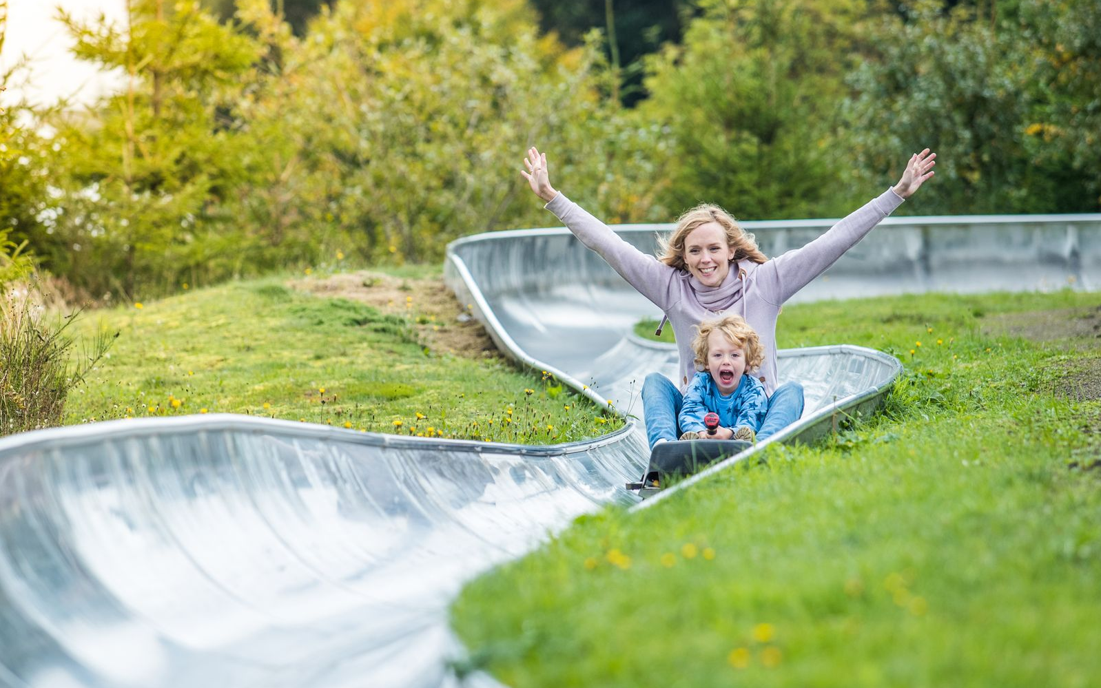 Eine Mutter fährt mit ihrem Kind auf dem Schoß die Sommerrodelbahn herunter. Das Kind jubelt vor Freude und die Mutter breitet die Arme aus.