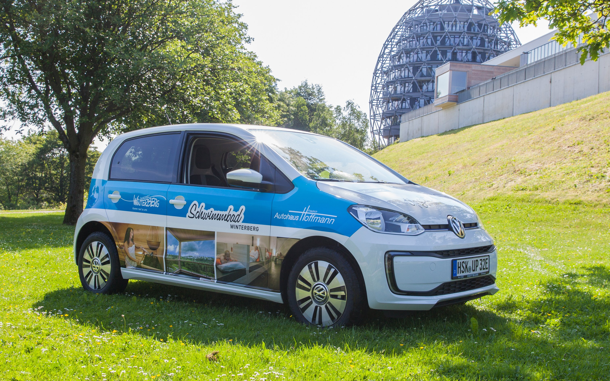 Ein Elektroauto der Marke VW Up auf einer Wiese mit Werbung für das Schwimmbad Winterberg.