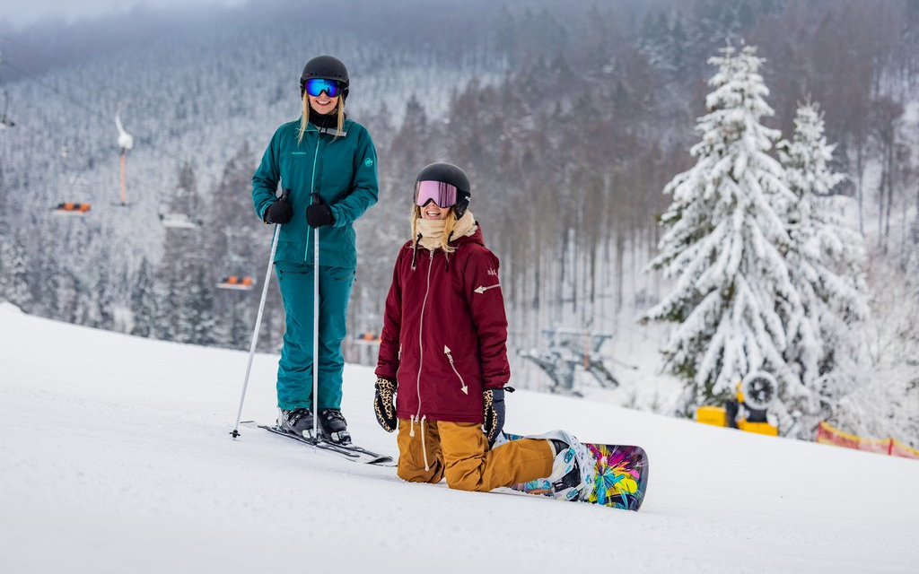 Zwei Frauen auf der Piste, eine mit Skiern, die andere mit Snowboard. Beide lächeln in die Kamera.