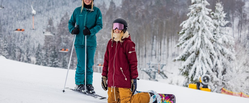 Zwei Frauen auf der Piste, eine mit Skiern, die andere mit Snowboard. Beide lächeln in die Kamera.