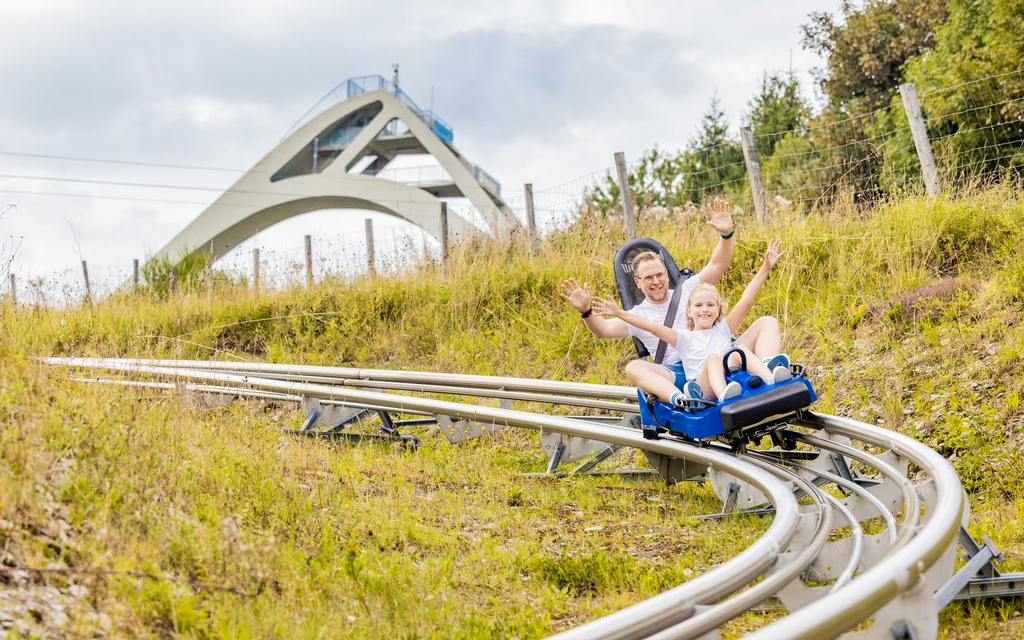 Ein Mann und ein Kind die auf einem Schlitten die Sommerrodelbahn herunterfahren. Im Hintergrund ist die St. Georg Sprungschanze zu sheen.