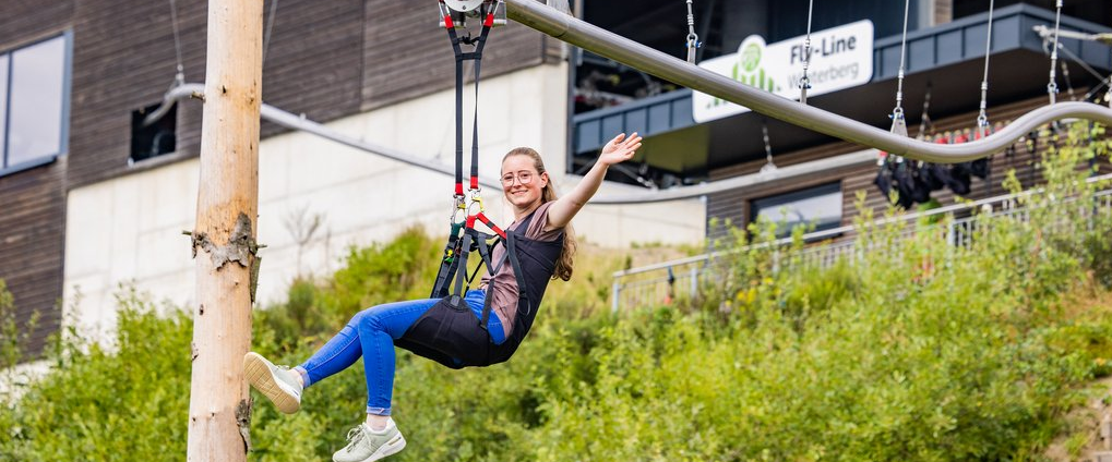 Eine Frau sitzt im Fluggeschirr in der Flyline und breitet lächelnd die Arme aus.