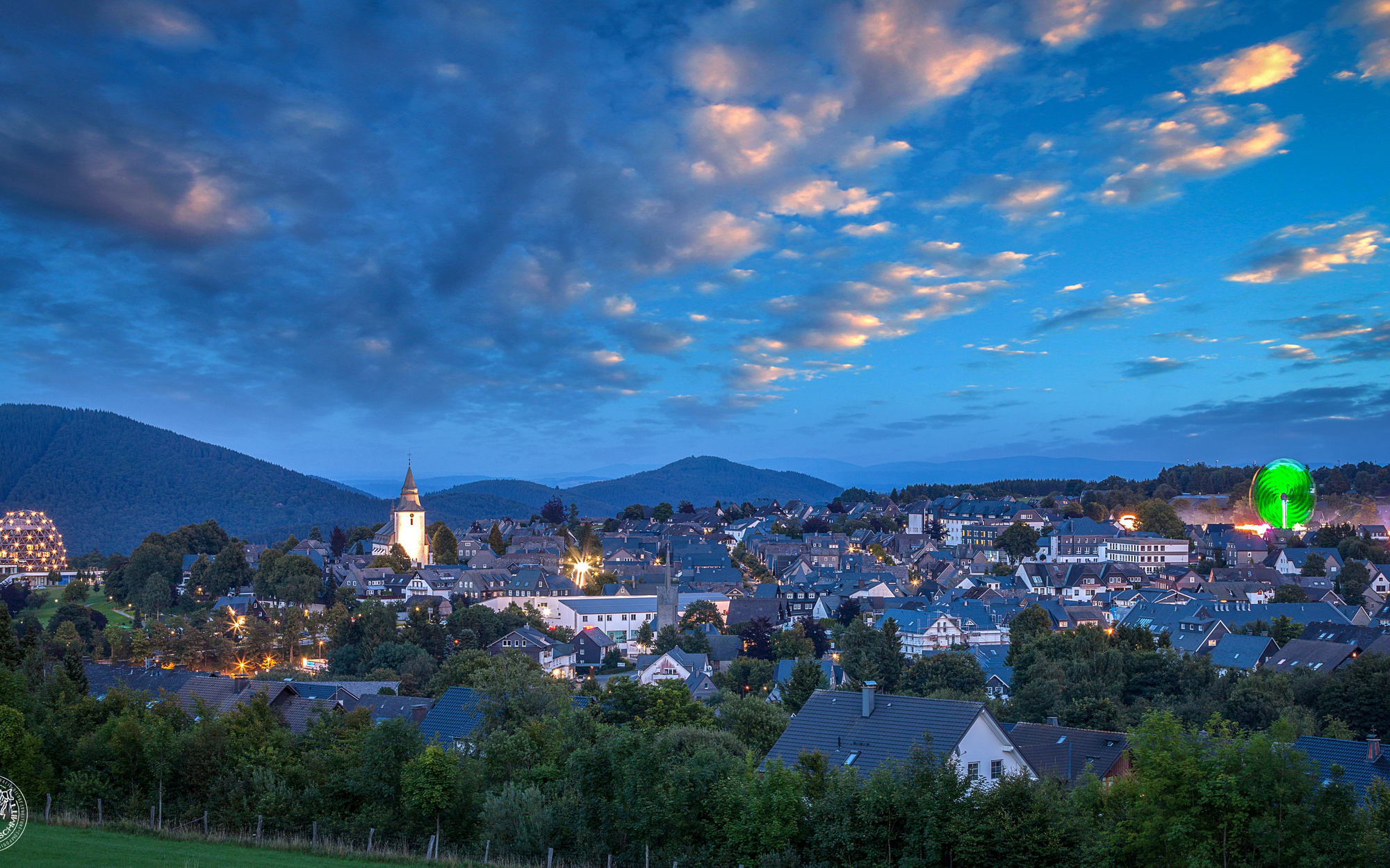 Aufnahme von einem Teil der Stadt Winterberg mit dem Hotel Oversum, der Kirche und einem Riesenrad auf der Kirmes in der Abenddämmerung.