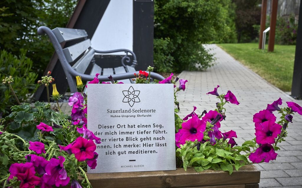 Ein Schild vor einer Bank mit der Aufschrift Sauerland Seelenorte Nuhne-Ursprung Erfurcht. Dieser Ort hat einen Sog, der mich immer tiefer führt, der vierte Blick geht nach innen. Ich merke: hier lässt sich gut meditieren.