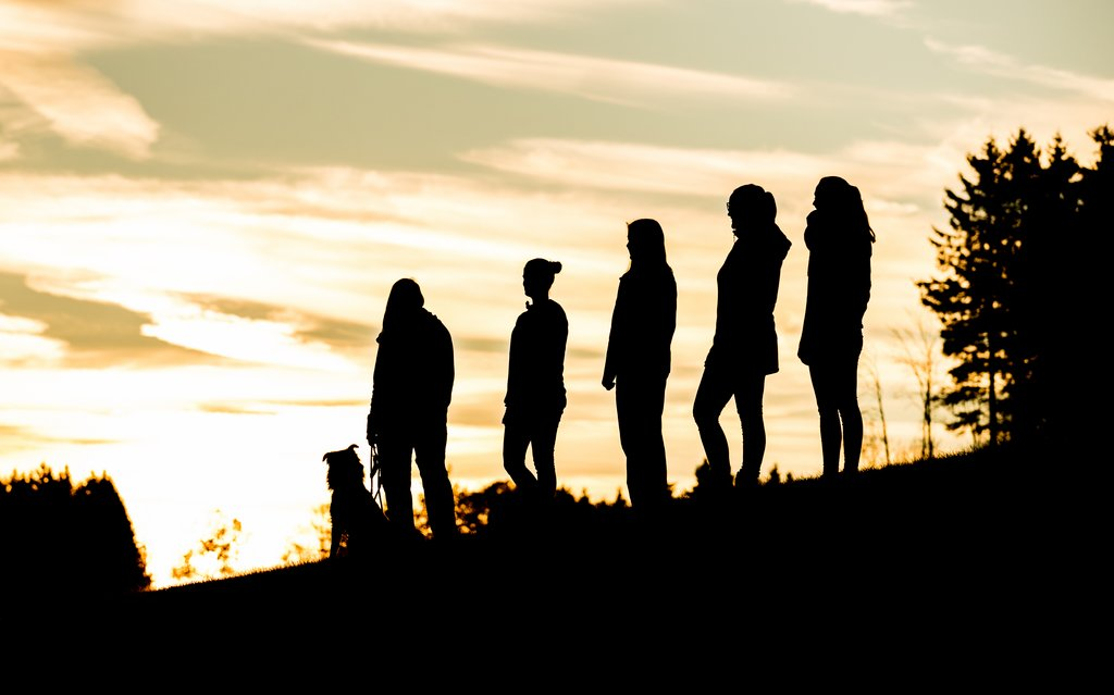 Fünf Frauen und ein Hund, die auf einer Wiese stehen und nur als dunkle Silhouetten zu erkennen sind.