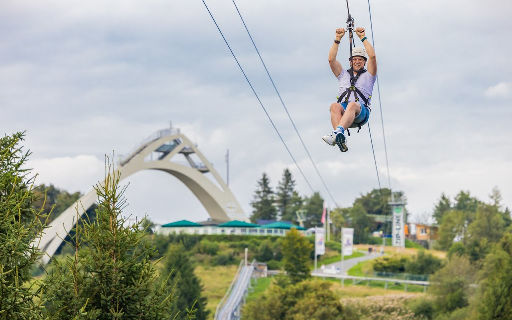 Ein Mann fliegt im Sitzen eine Zipline herunter. Im Hintergrund ist die St. Georg Sprungschanze zu sehen.