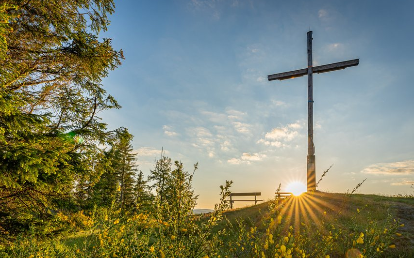Das Gipfelkreuz auf dem Alten Grimmen mit Bank und aufgehender Sonne im Hintergrund.