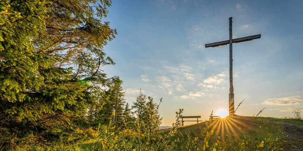 Das Gipfelkreuz auf dem Alten Grimmen mit Bank und aufgehender Sonne im Hintergrund.