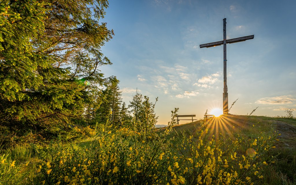 Das Gipfelkreuz auf dem Alten Grimmen mit Bank und aufgehender Sonne im Hintergrund.