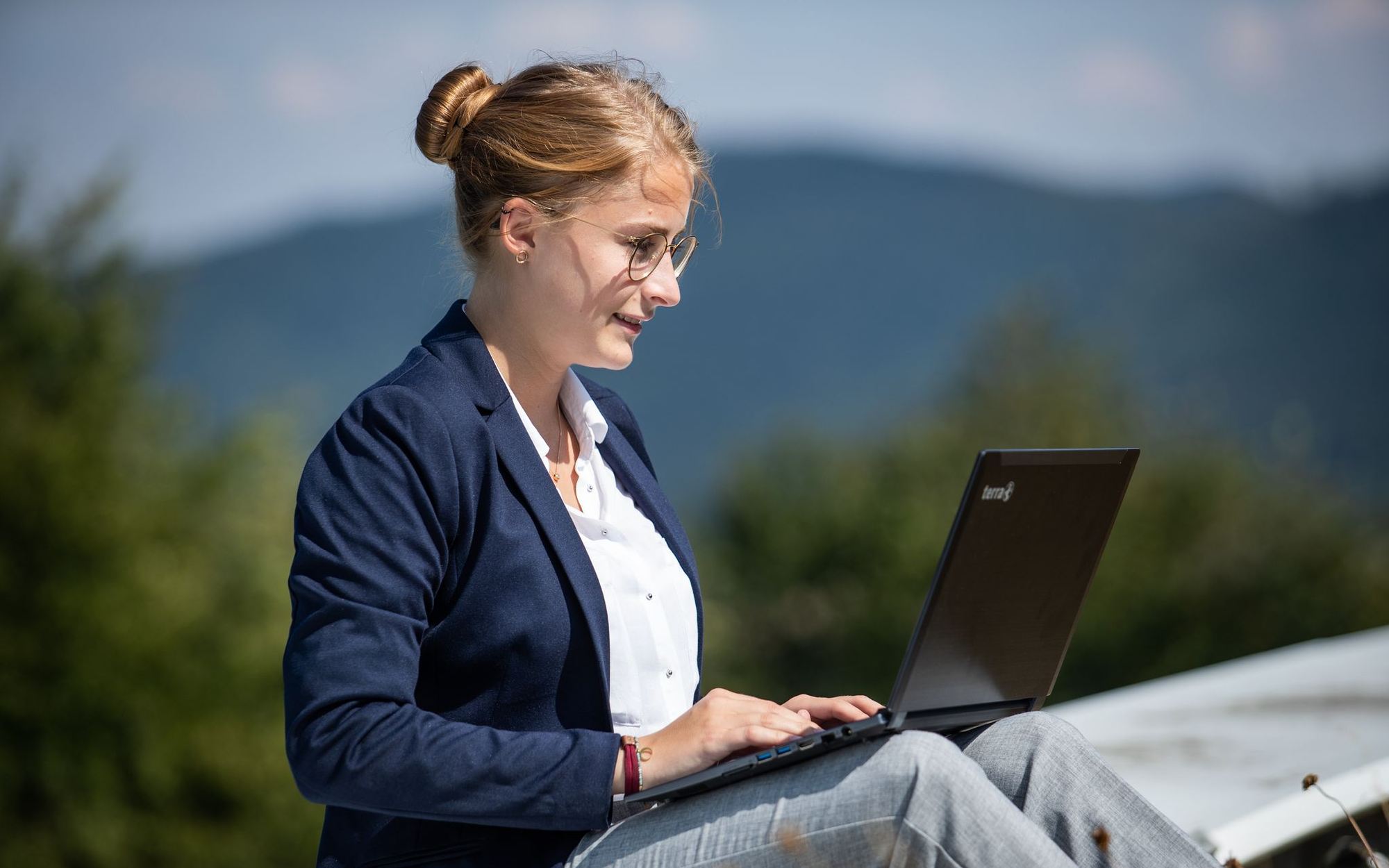 Eine Frau sitzt mit ihrem Laptop auf den Knien im Freien und arbeitet.