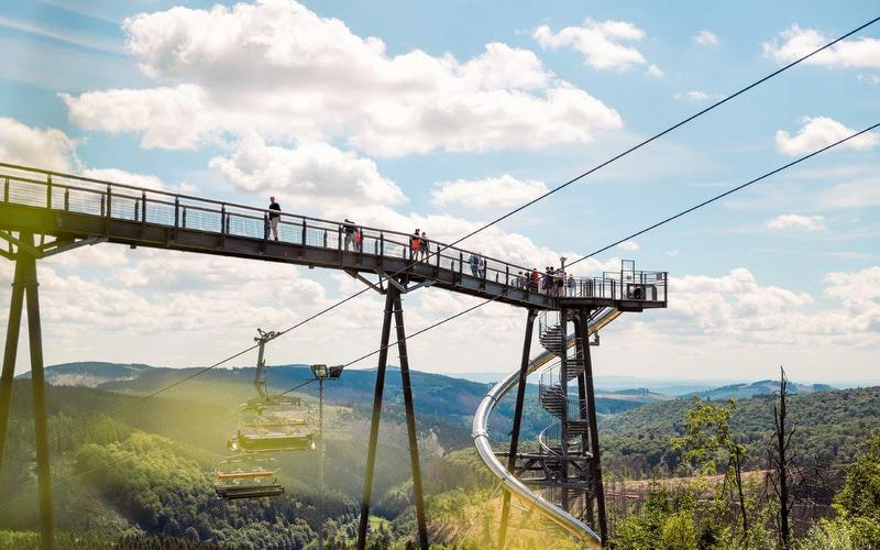 Mehrere Personen stehen am Ende der Panorama Erlebnisbrücke. Unter ihnen fährt ein Skilift nach oben und am Ende der Brücke sind eine Rutsche und eine Wendeltreppe zu sehen.