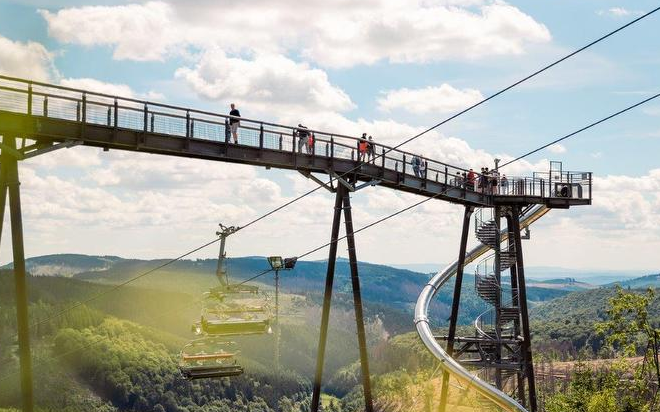 Mehrere Personen stehen am Ende der Panorama Erlebnisbrücke. Unter ihnen fährt ein Skilift nach oben und am Ende der Brücke sind eine Rutsche und eine Wendeltreppe zu sehen.
