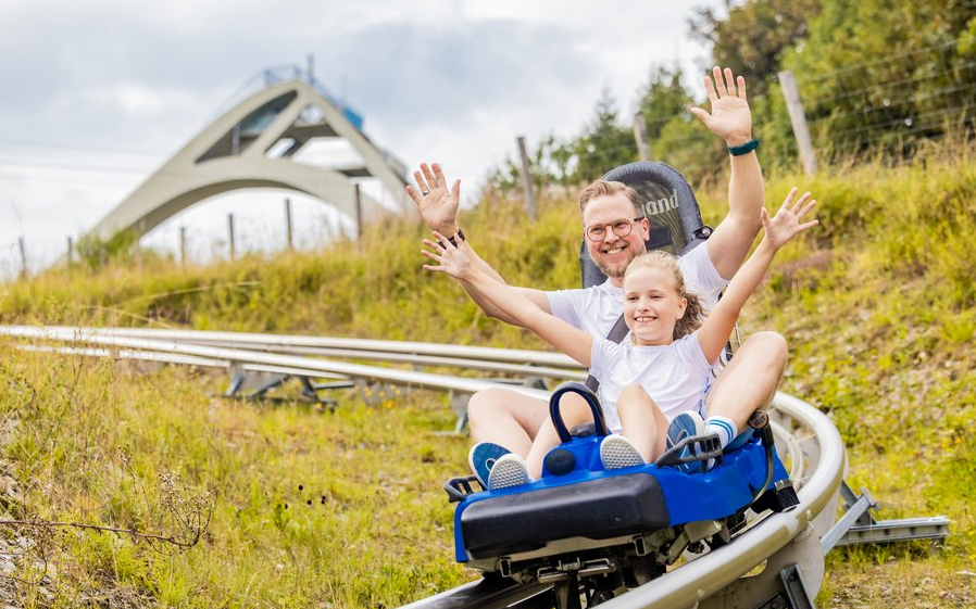 Ein Mann und ein Kind fahren mit ausgebreiteten Armen auf einem Schlitten die Sommerrodelbahn im Vordergrund der St. Georg Sprungschaze herunter.