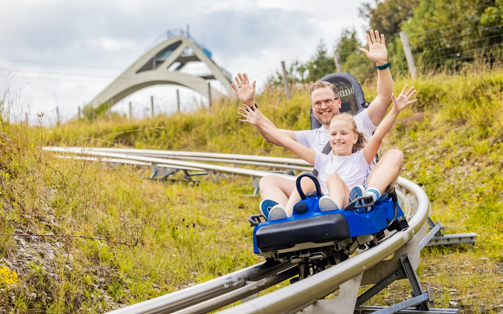Ein Mann und ein Kind fahren mit ausgebreiteten Armen auf einem Schlitten die Sommerrodelbahn im Vordergrund der St. Georg Sprungschaze herunter.