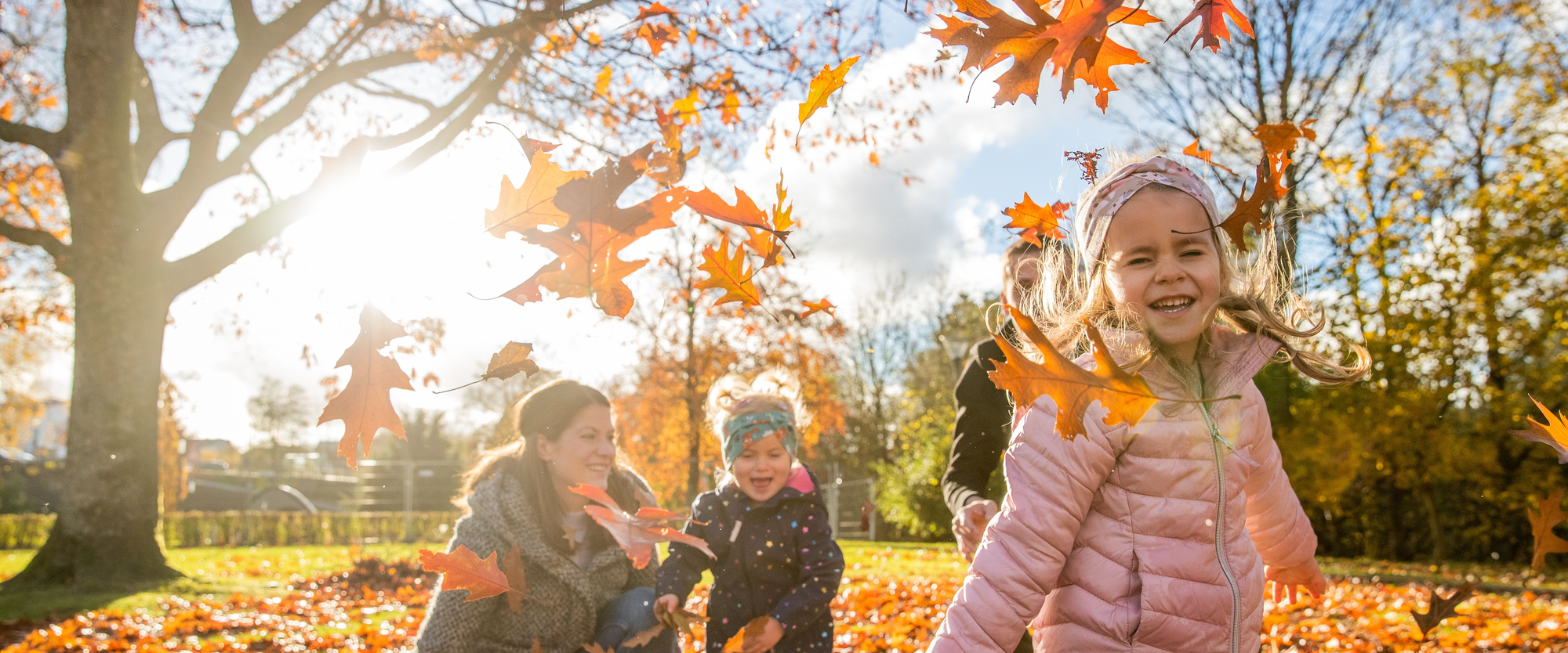 Eine Familie mit zwei Kindern spielt draußen im Sonnenschein und wirft bunte Herbstblätter in die Luft.