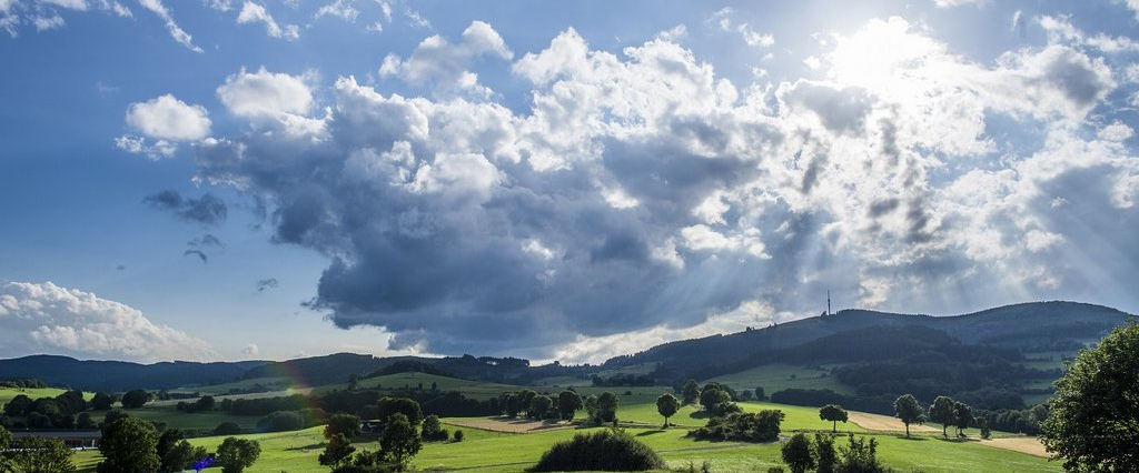 Landschaftsfoto mit weiten grünen Wiesen, blauem Himmel mit Wolken und Bergen im Hintergrund.